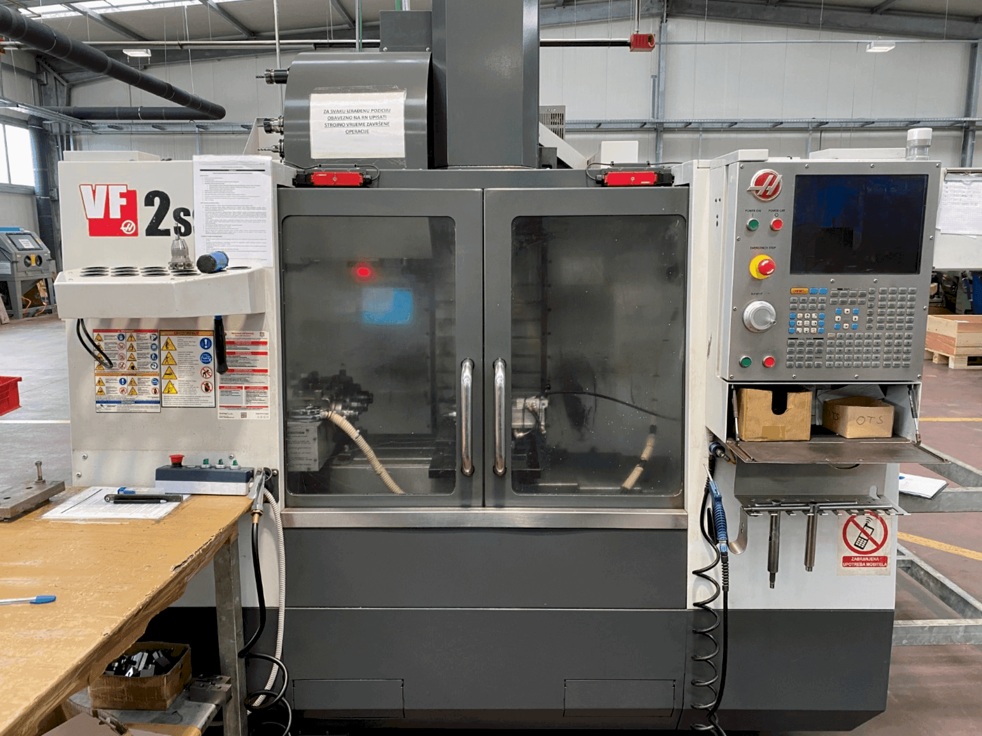 Haas VF-2SS töötluskeskus esiplaanil, millel on juhtpaneel, jahutusvedeliku voolik ja ohutussildid küljel.